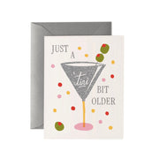 A greeting card with a gray envelope behind it, featuring a martini glass illustration and the text 'JUST A TINI BIT OLDER' with colorful confetti dots and olives on a white card.