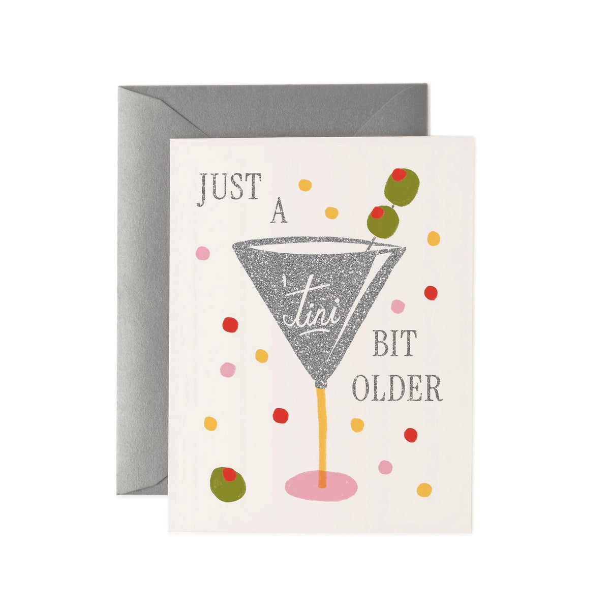 A greeting card with a gray envelope behind it, featuring a martini glass illustration and the text 'JUST A TINI BIT OLDER' with colorful confetti dots and olives on a white card.