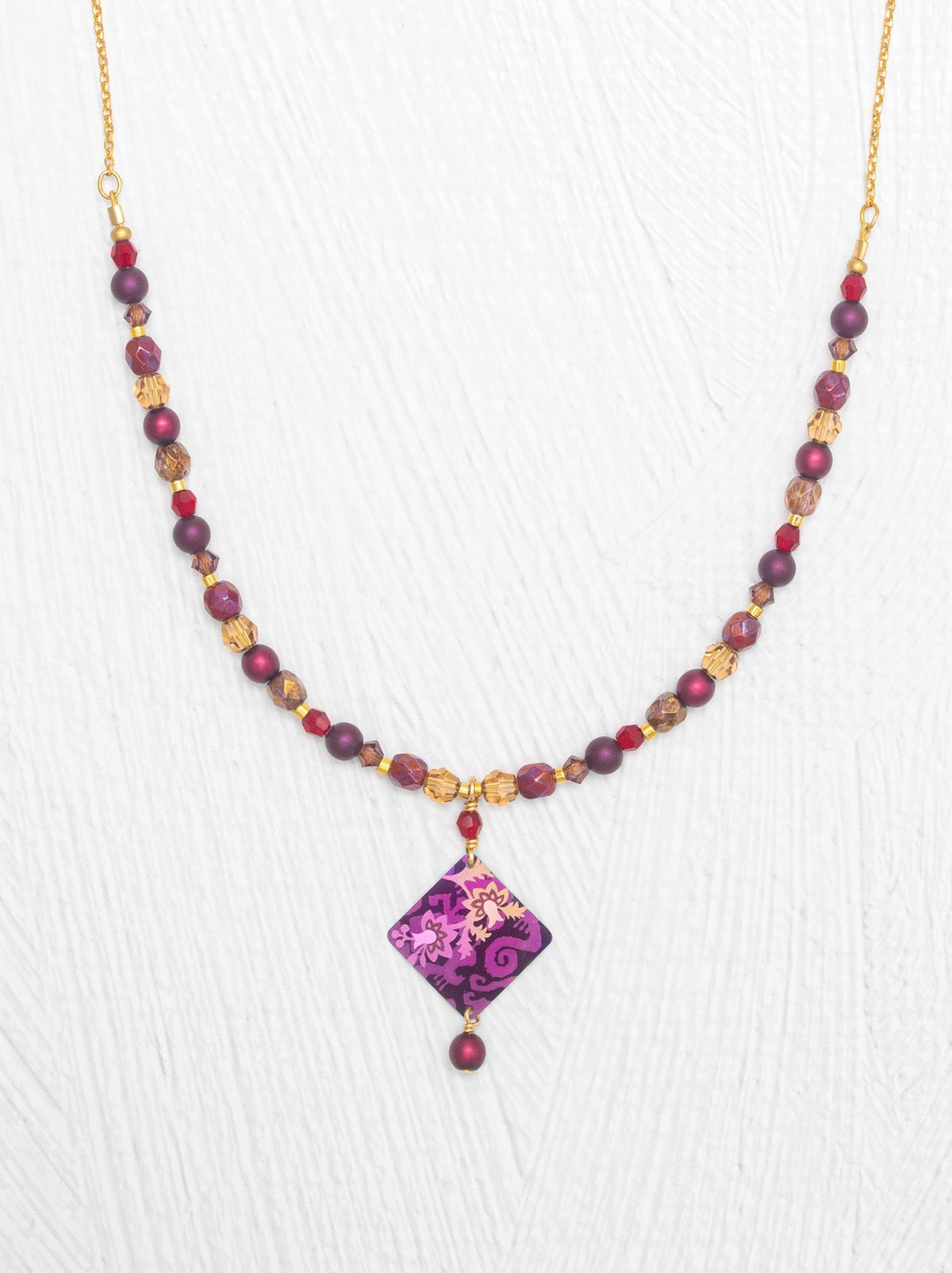 A beaded necklace featuring faceted purple and red beads, gold-toned spacers, and a central diamond-shaped purple pendant with a floral pattern, displayed on a white textured background.