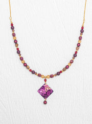 A beaded necklace featuring faceted purple and red beads, gold-toned spacers, and a central diamond-shaped purple pendant with a floral pattern, displayed on a white textured background.