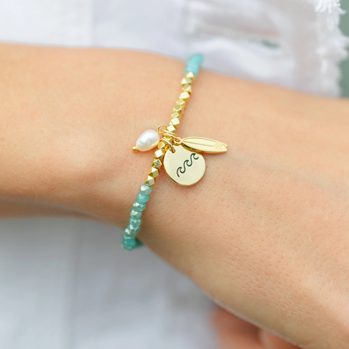 A close-up of a person's wrist wearing a delicate gold charm bracelet with a turquoise beaded cord, a white pearl, and a gold disc engraved with the zodiac symbol Aquarius.